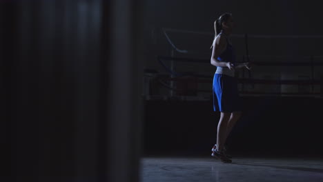 Entrenamiento-Cardiovascular-Para-Luchadores.-Una-Joven-Y-Hermosa-Boxeadora-Profesional-En-Entrenamiento-En-El-Pasillo-Salta-Sobre-Una-Cuerda-A-La-Luz-De-La-Contra-En-Una-Habitación-Oscura-Con-Ropa-Azul