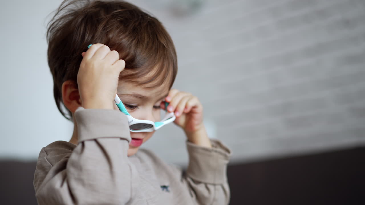 Lovely Caucasian boy wearing grey sweater puts on sunglasses. Cropped image of a kid indoors.