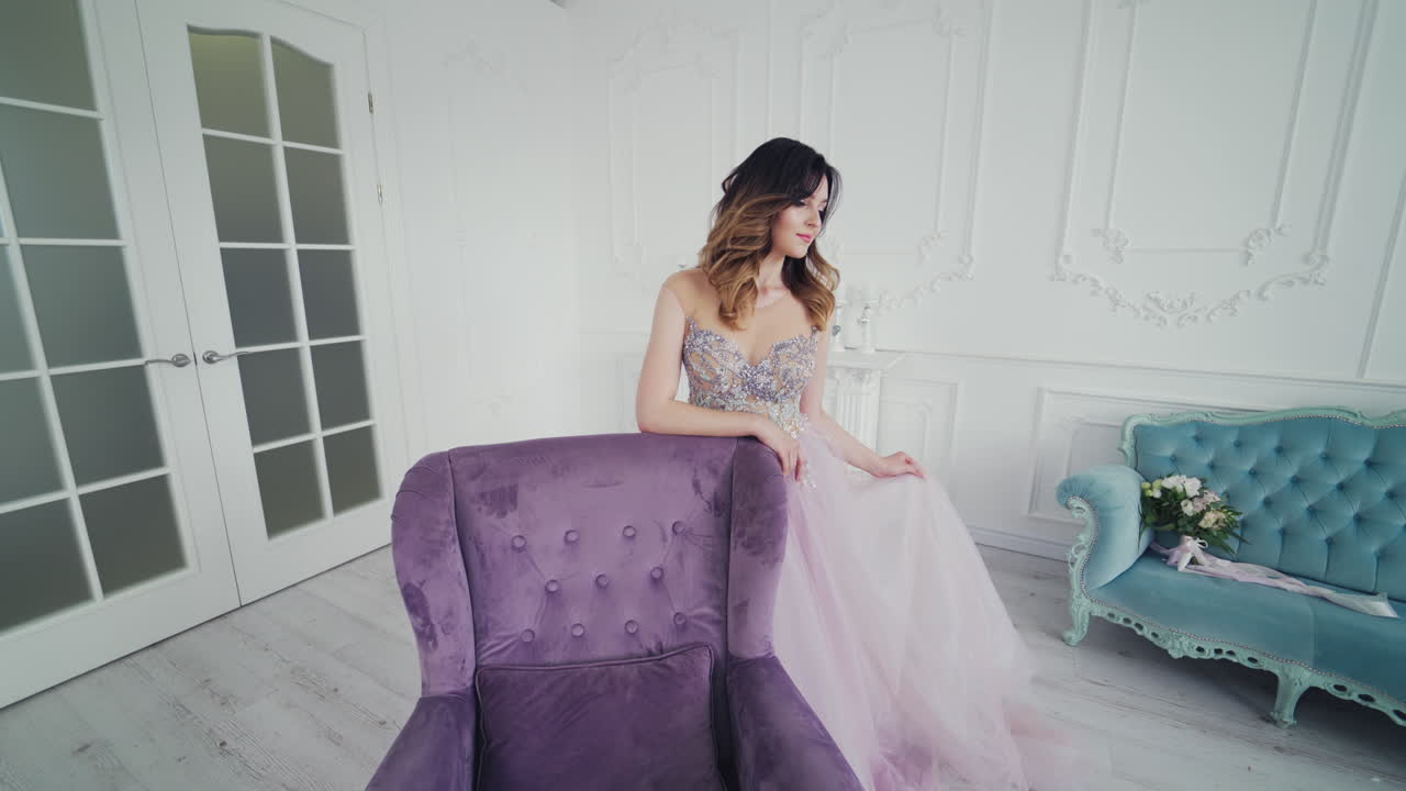 Fantastic young woman in a luxury dress is posing to camera. Beautiful model is standing behind the royal armchair in a long wedding dress on the background of studio.
