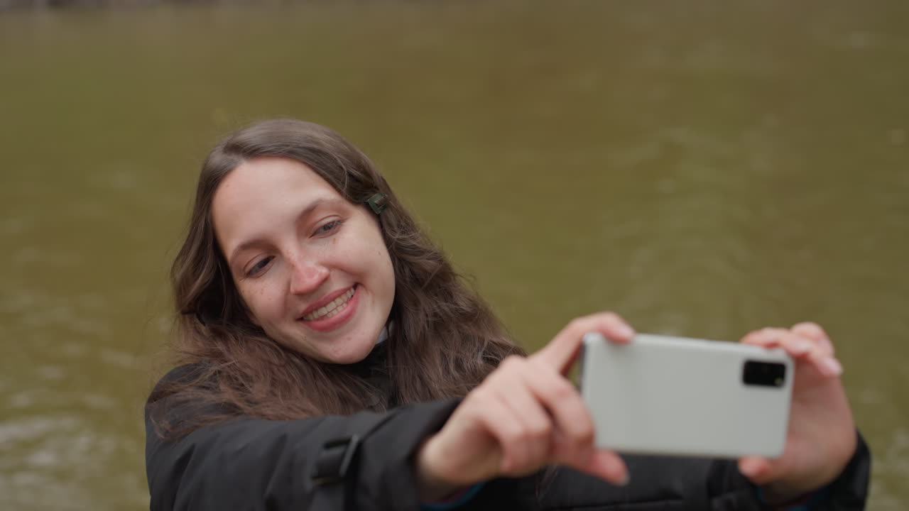 Mulher perto da água. Mulher contemplativa em pé ao lado de um riacho calmo. Mulher de cabelos longos tira uma foto perto de água tranquila. Mulher pensativa de casaco escuro captura sua imagem ao lado de um rio de fluxo suave