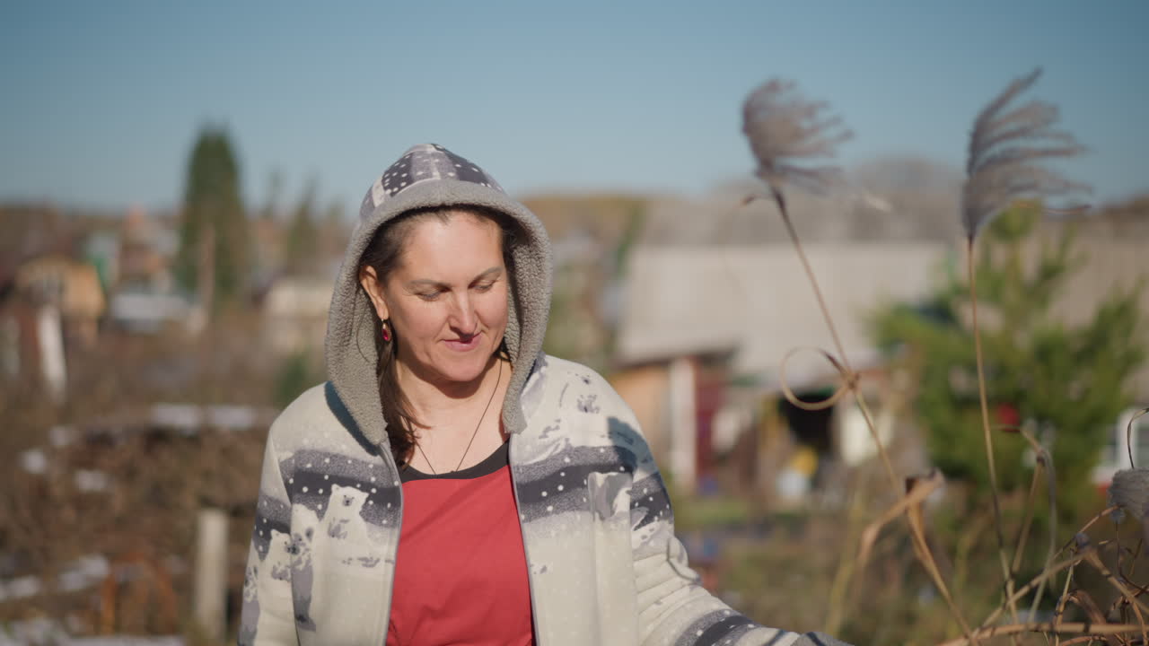 Lächelnde Frau im Hausgarten mit Kapuzenpullover. Ein sonniger Garten mit Pflanzen und kleinen Häusern im Hintergrund. Ihr Ausdruck ist lässig und warm, während sie das Laub berührt. Ein offenes, freundliches Porträt mit gemütlicher Herbststimmung.