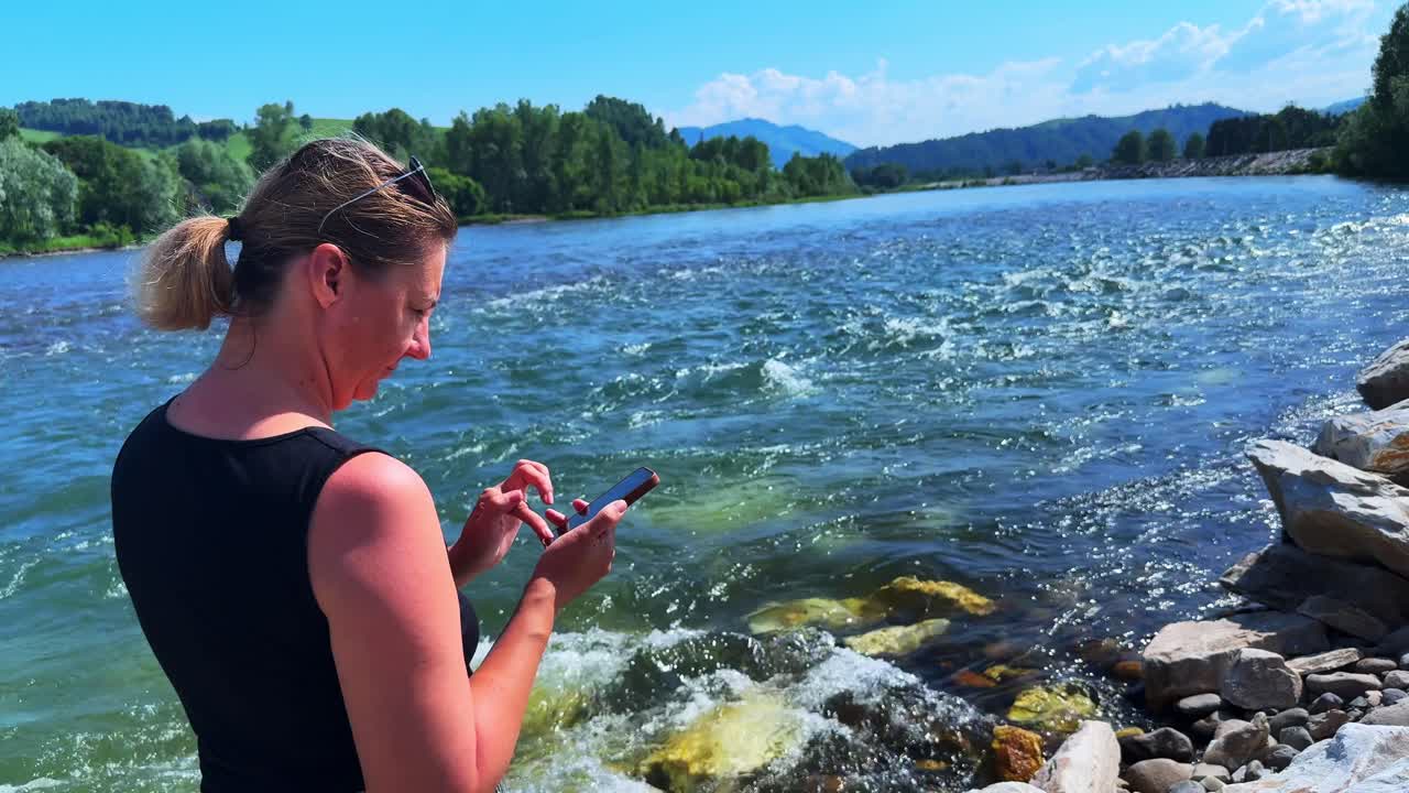 A Woman Engaged in Communication by the Riverbank: Disconnecting from Nature to Connect with Technology While Enjoying a Beautiful Scenic View