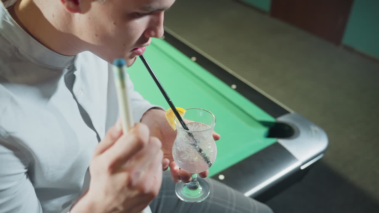 색 셔츠와 회색 바지를 입은 젊은 남자의 클로즈업, 녹색 빌리어드 테이블에 앉아 큐 스틱과 검은 을 가진 레몬 음료 한 잔을 들고.