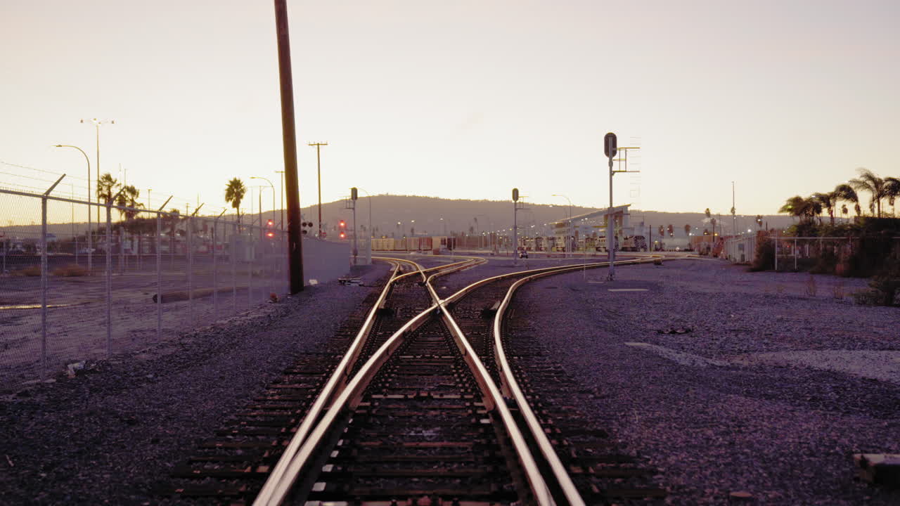 Slowly moving down an empty railway while approaching a fork. A mountain in the background is lit with a golden tint. POV