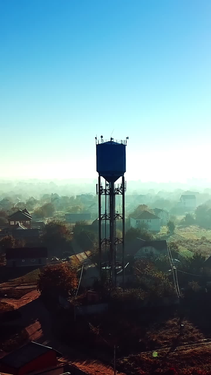 Birds-eye view of a beautiful morning landscape of a rural place. Flying over the small houses with gardens and high construction at sunrise. Aerial view. Camera moves forward. Slow motion. Vertical video
