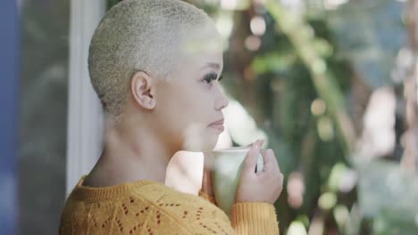 Happy-biracial-woman-drinking-coffee-at-window-at-home-in-slow-motion