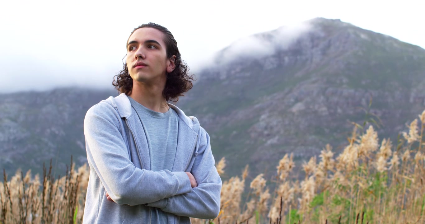 Hombre afroamericano de unos 20 años de pie con sudadera con capucha y brazos cruzados entre la hierba, observando las nubes.