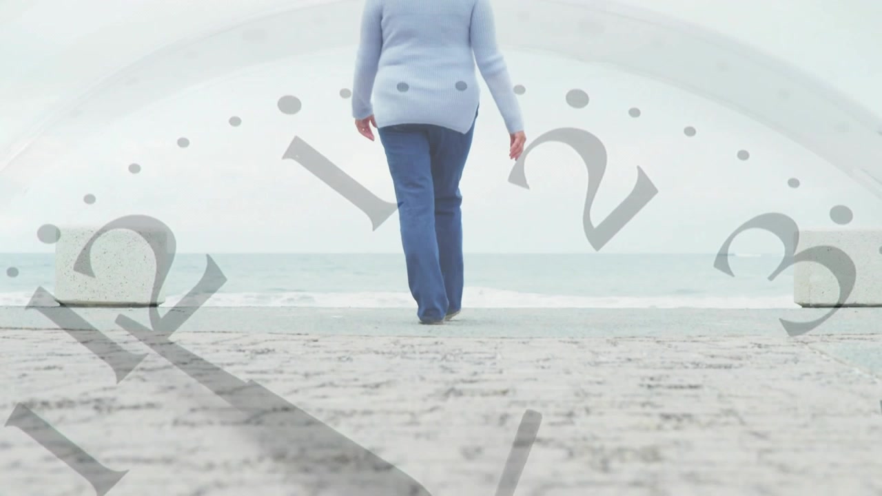 Low camera rising revealing senior woman walking to sea while clock hands shifting marking time