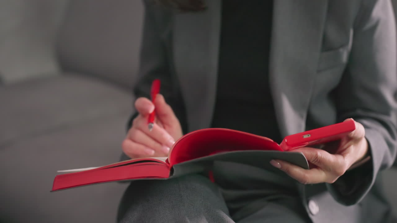 Businesswoman holding red notebook, focusing on reading and writing, modern office setting, professional attire, sitting relaxed on couch, planning, thinking, productivity, decision-making