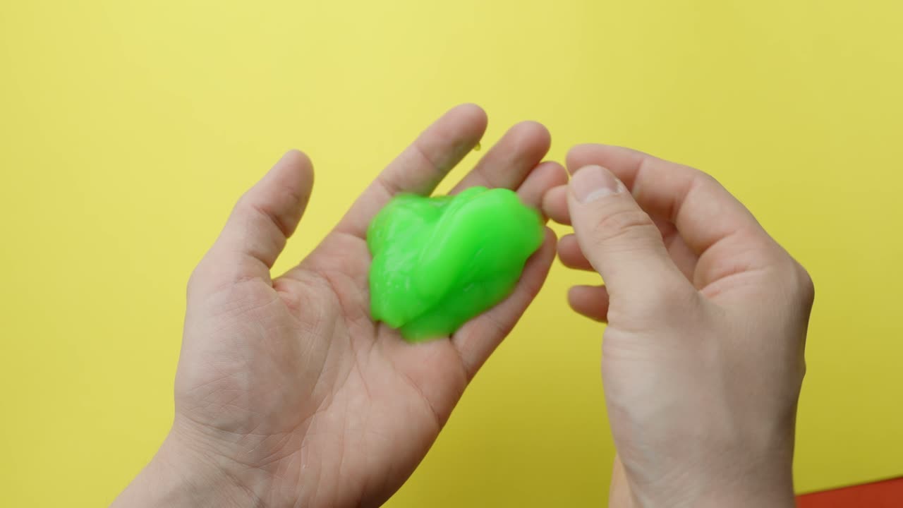 Hands stretching and pressing a wavy green slime toy for ASMR and stress relief against a yellow background, highlighting texture and movement, static camera, real time, close-up view