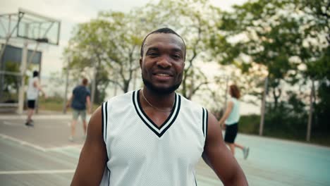 A-Black-person-in-a-white-T-shirt-poses-and-smiles-on-the-basketball-court-in-front-of-his-friends-who-are-playing-basketball