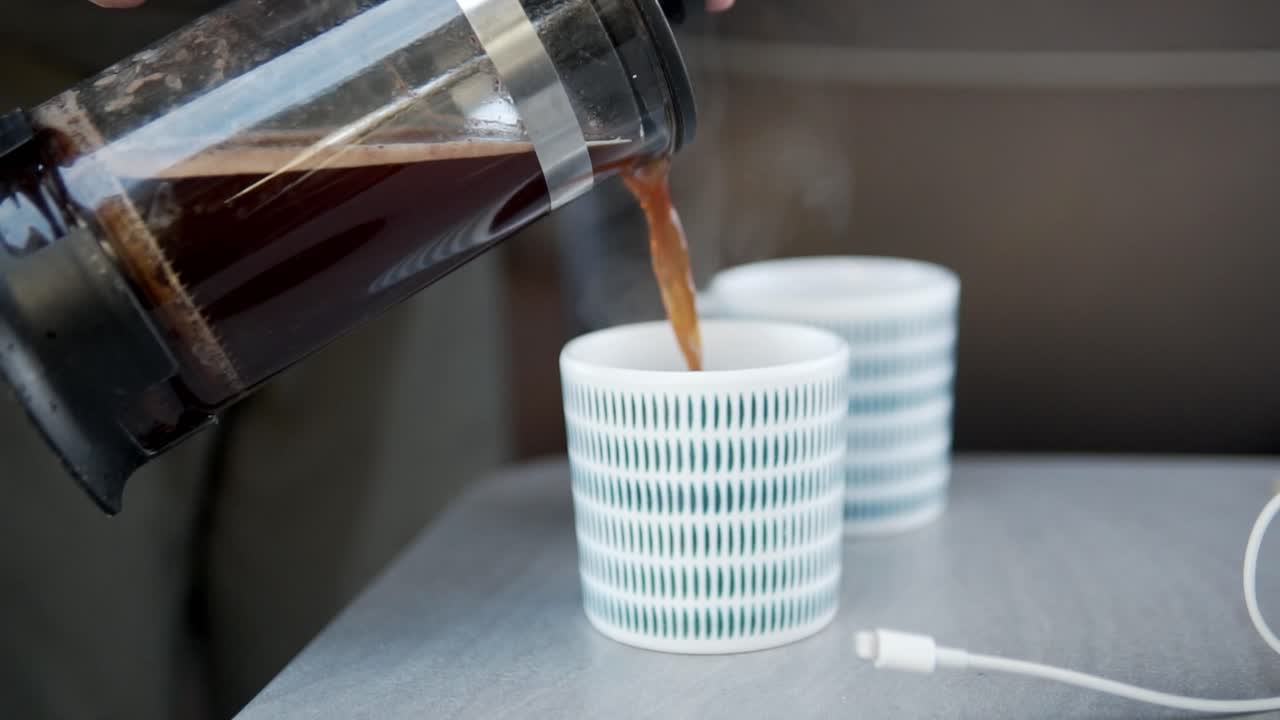 A close-up shot of rich, dark coffee being poured from a clear glass French press into two textured cups, highlighting the comforting ritual of serving a hot beverage