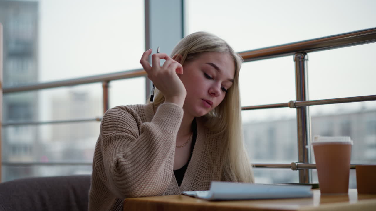 созерцательный студент сидит за столом, пишут задумчиво с чашечкой чая перед ней и размытым видом зданий на заднем плане, создавая мирную и увлекательную атмосферу в кафе
