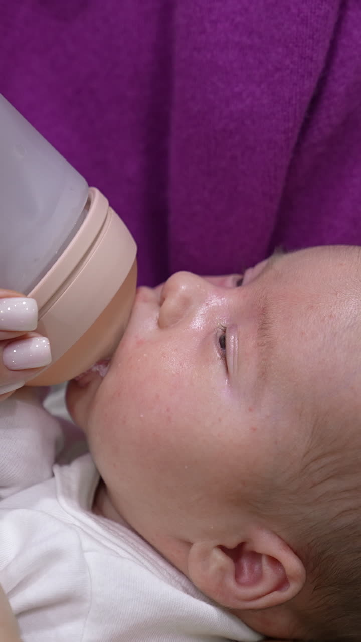 Sweet little baby eating milk from a bottle in mommy's hands. Baby finishing the bottle and lets go the bottle pacifier. Suckling baby close up. Vertical video