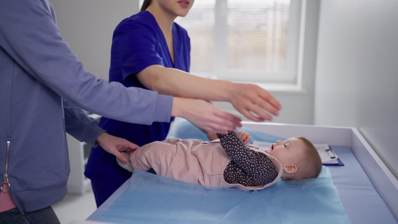 A confident and happy brunette mother and her daughter came to the childrens pediatrician for preventive care. Little girl baby lies in front of girl pediatrician doctor in modern clinic