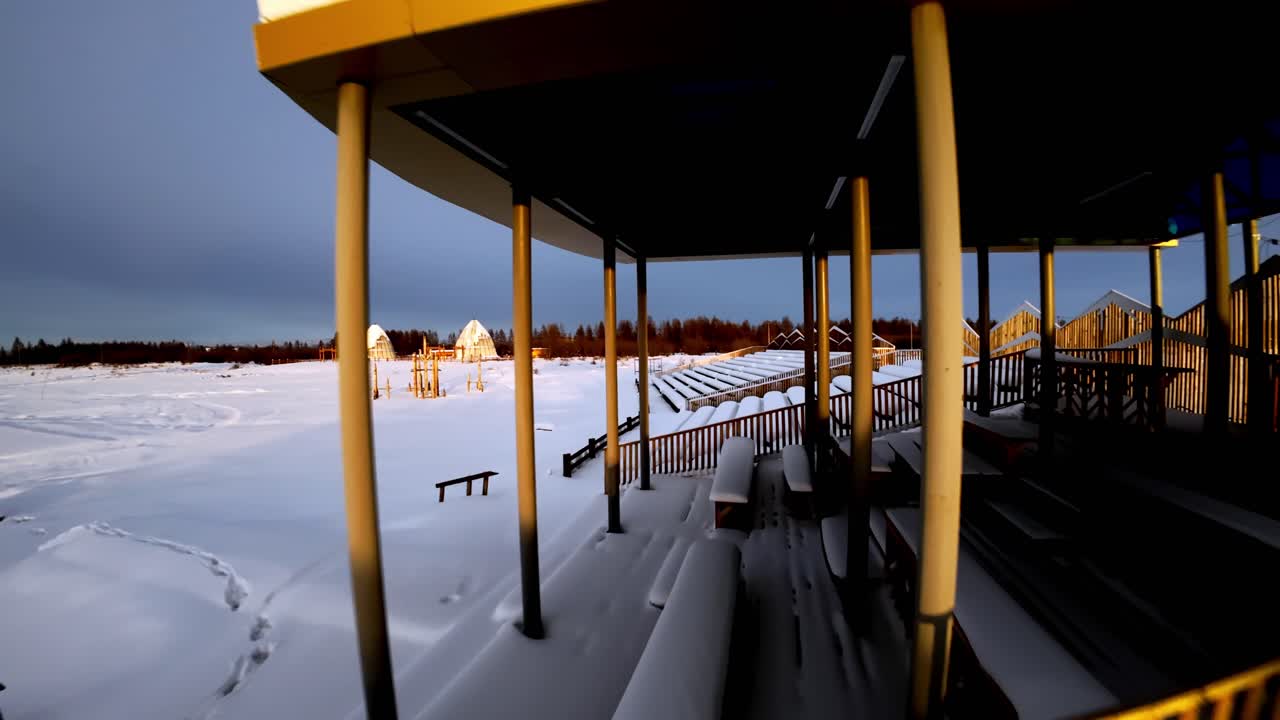 Ein sanfter Drohnenflug gleitet über schneebedeckte Holztribünen in einer ruhigen Winterlandschaft. Warmes Sonnenlicht hebt die Strukturen hervor und schafft eine ruhige und filmreife Szene.