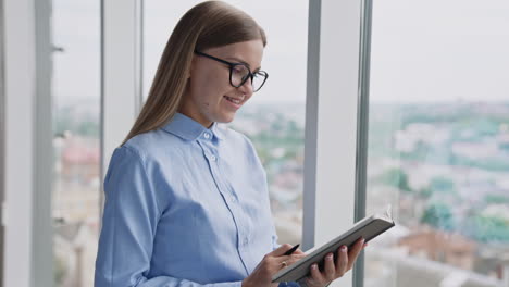happy positive smiling woman standing at the big panoramic window. businesswoman taking notes into paper book and then looking into window. cityscape at backdrop in blur.