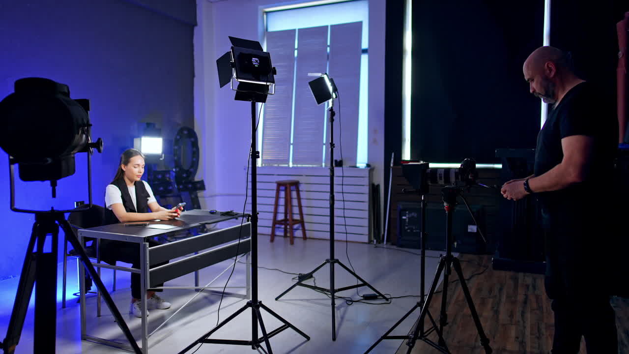 Interior of the modern photo studio with necessary equipment. Smiling lady sits at desk. Cameraman prepares light for footage.