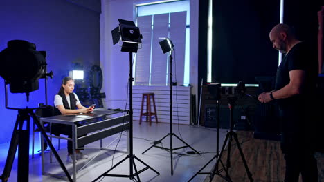 interior of the modern photo studio with necessary equipment. smiling lady sits at desk. cameraman prepares light for footage.
