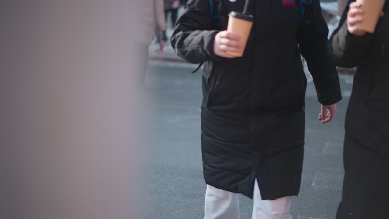 low angle street view shows pedestrians in winter coats carrying hot coffee, boots stepping from curb onto wet asphalt near crosswalk, cold urban morning commute with crowd moving toward sidewalk
