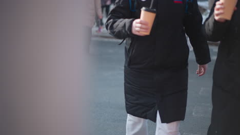 low angle street view shows pedestrians in winter coats carrying hot coffee, boots stepping from curb onto wet asphalt near crosswalk, cold urban morning commute with crowd moving toward sidewalk