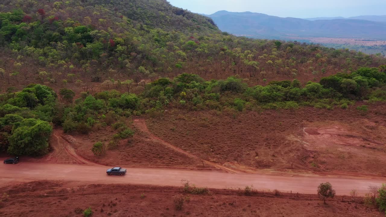 Un drone si muove lateralmente seguendo un pickup grigio 4x4 a doppia cabina mentre percorre una strada sterrata rossa dove gli alberi fitti del Cerrado incontrano la boscaglia aperta sulla strada per Tocantins