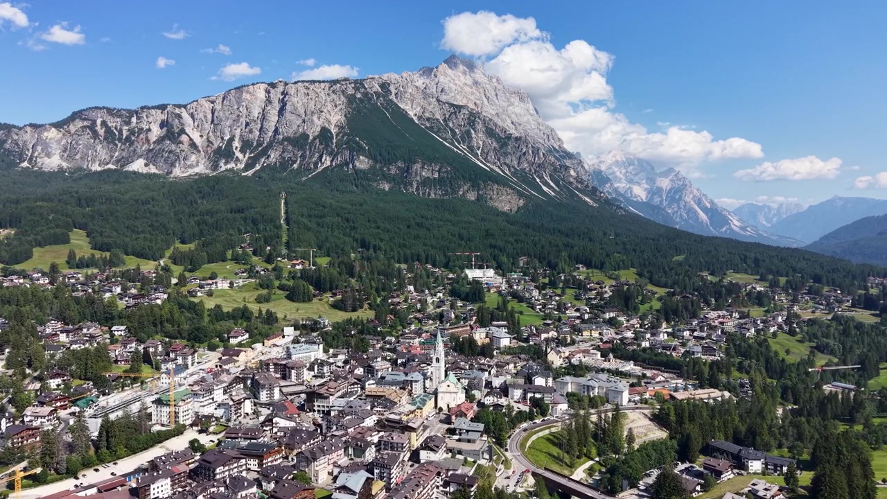 Luftaufnahme mit Drohne über Cortina d'Ampezzo, mit der Stadt und der umliegenden Alpenlandschaft, eingerahmt vom dramatischen Kalksteinmassiv des Berges Pomagagnon unter einem klaren Sommerhimmel
