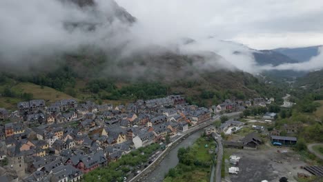 the drone initiates with a view of the quaint spanish town in the pyrenees mountains and then smoothly pans to unveil the cloudy, majestic mountain landscape of the pyrenees