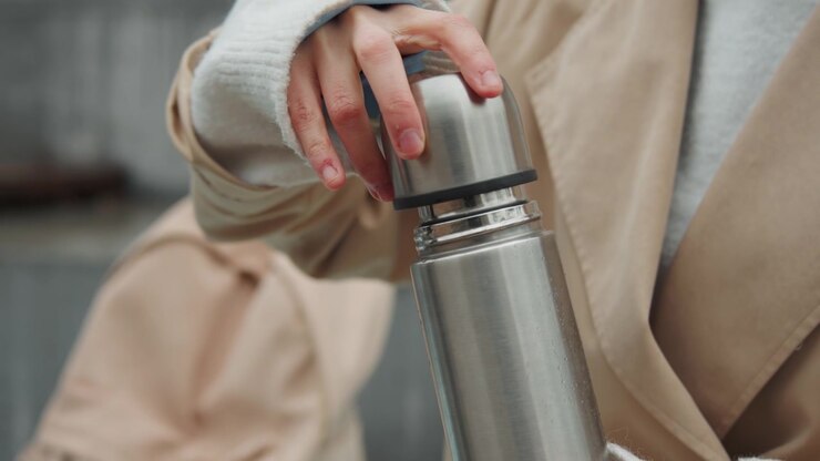 Close up of woman in beige coat and white sweater holding stainless steel thermos unscrewing lid to drink. Thermos with condensation visible on metal surface. Out of focus urban background