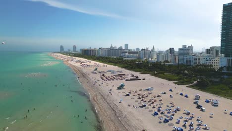 aerial 4k view of miami beach, florida, as the drone soars above the coastline, revealing a lively shore filled with beachgoers, colorful umbrellas, and waves rolling in under the bright tropical sky.