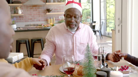 happy african american grandfather holding hands with family saying grace at christmas dinner table