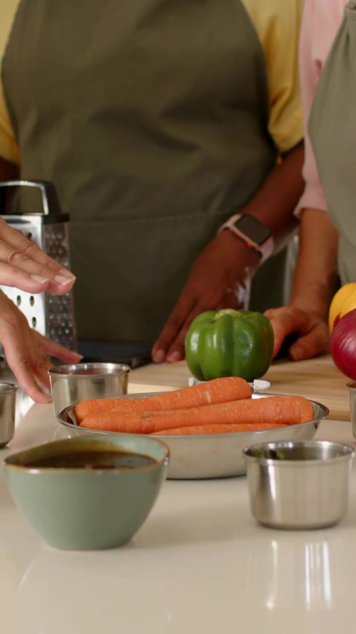 Video verticale: Una donna anziana con un grembiule verde legge un tablet mentre sistema verdure e tazze sul bancone per cucinare.