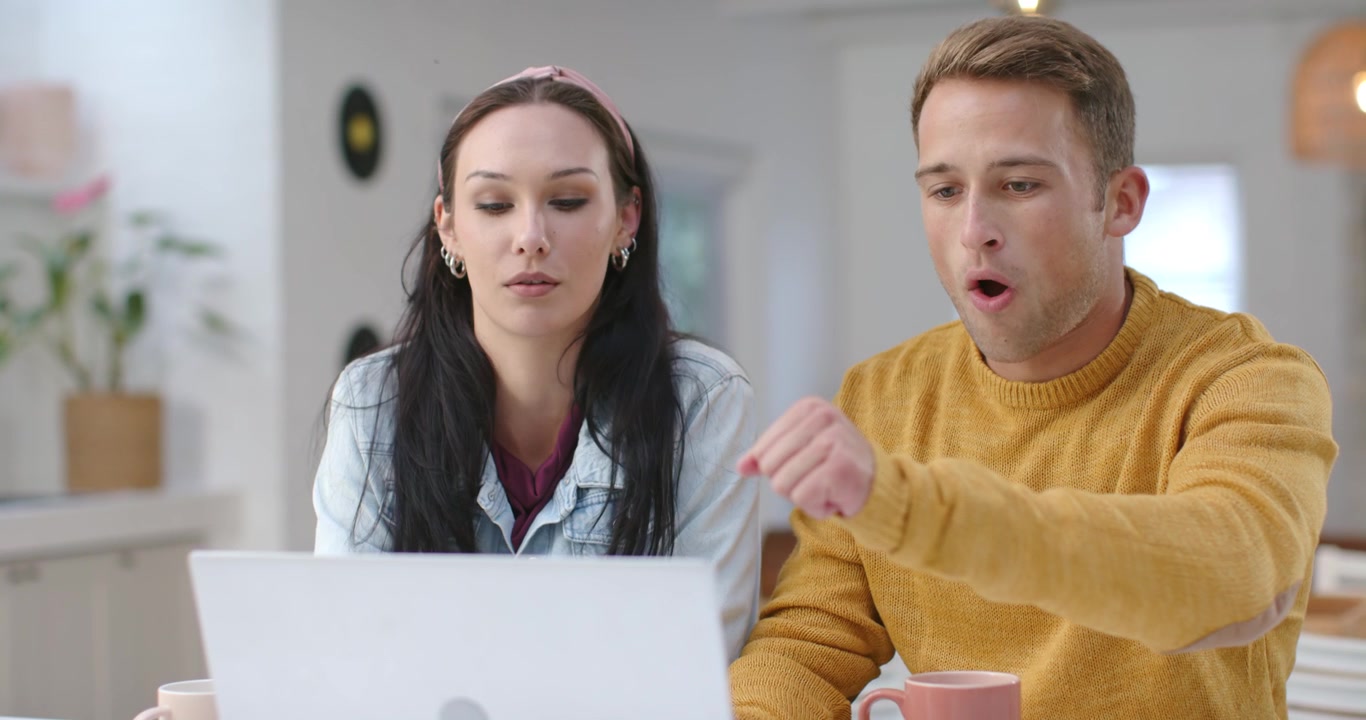 Couple in 20s sitting at kitchen table, woman showing card while man typing laptop to pay