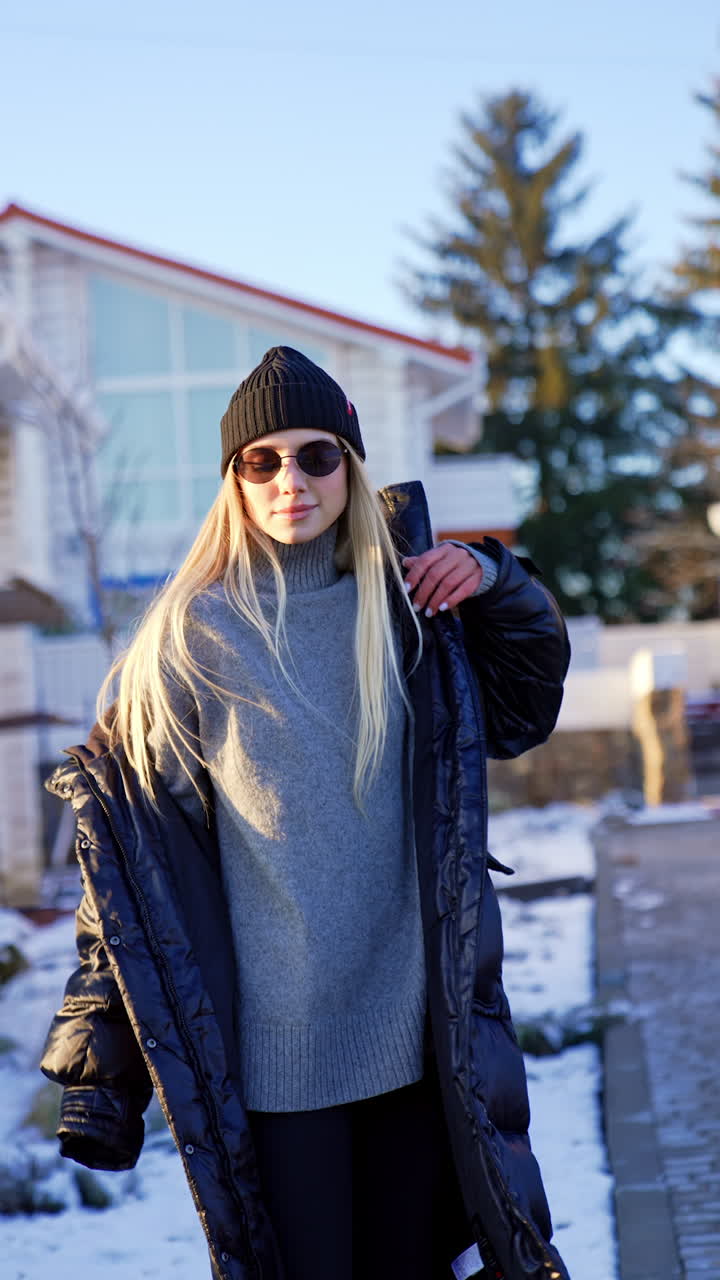 Blonde girl wearing black cap, coat and leggings, sunglasses and grey sweater poses outdoors. Winter season fashion. Vertical video.