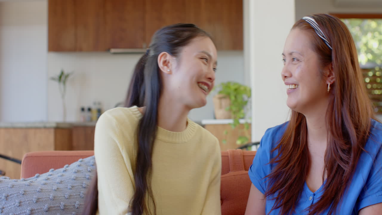 Smiling and talking, mature asian mother and daughter bonding on couch at home