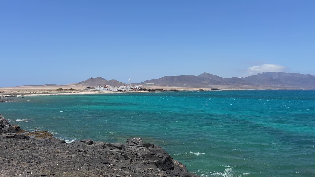Scenic view from the rocky shore of the tiny fishing village El Puertito de la Cruz, with its white houses and calm, turquoise bay. Jandia, Fuerteventura, Spain