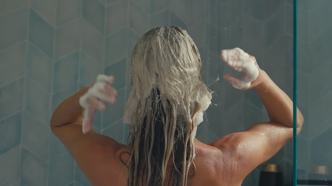 Una donna bionda si lava i capelli sotto la doccia, massaggiando lo shampoo schiumoso mentre l'acqua le scorre addosso