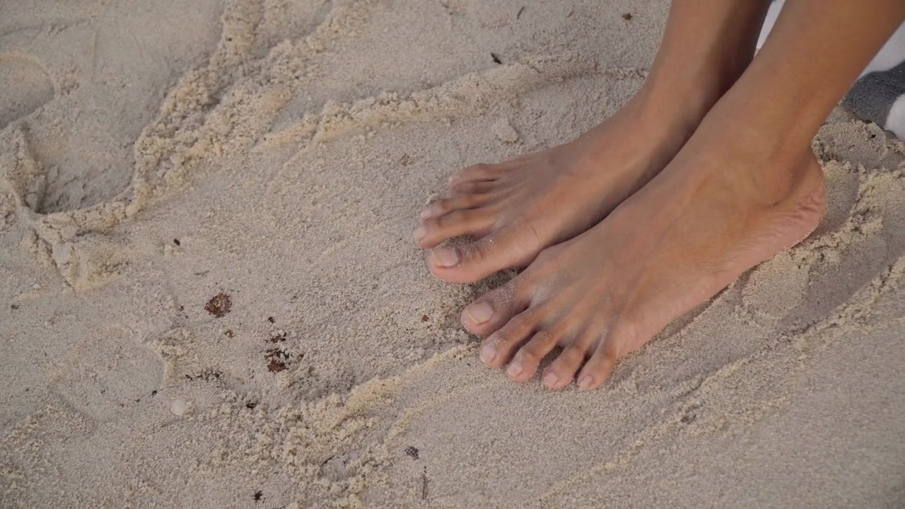 Sandali boho con conchiglie sulla sabbia, primo piano di piedi che tolgono calzature ornate di conchiglie, granelli delicati tra le dita dei piedi, luce dorata, cavigliera e dettagli di gioielli, brezza leggera, pedicure e atmosfera vacanziera, sabbia tattile.