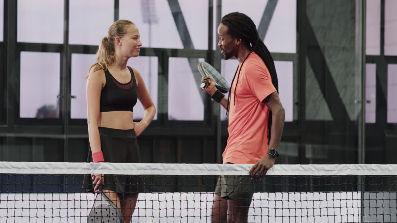 Smiling diverse athletes discussing padel tennis strategy on court, holding rackets