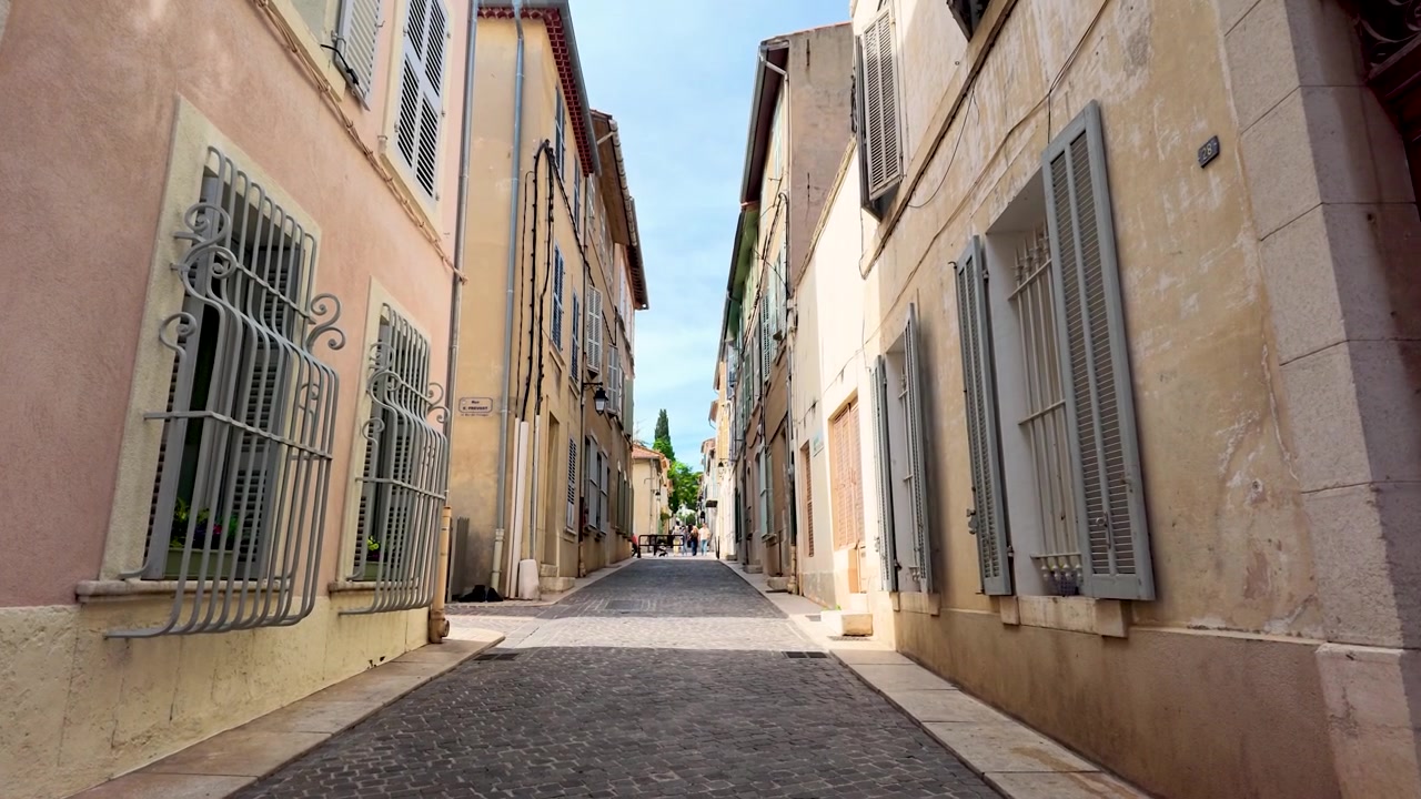 Ripresa in movimento dal basso attraverso una stretta strada acciottolata fiancheggiata da edifici mediterranei color pastello.