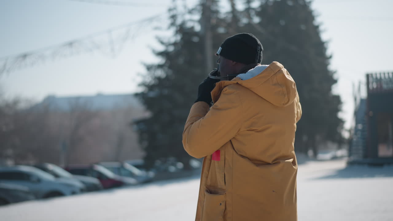 round view of shutterbug in yellow parka and beanie capturing snowy urban landscape with vintage camera, strap on neck, cars and buildings blurred in background under soft cold winter daylight