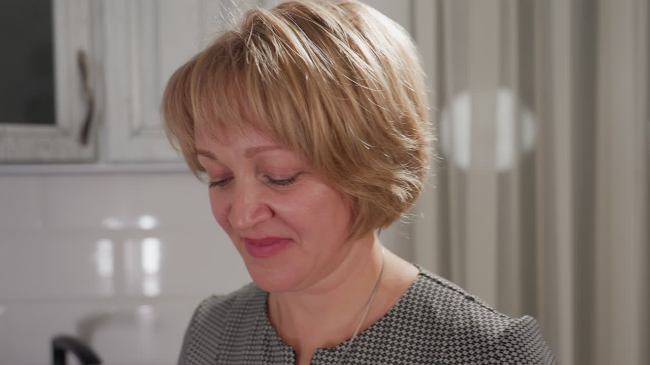 Close up of female artist smiling softly while making gentle body movements as she works joyfully in kitchen with light reflecting on tiled wall and faint curtain patterns in background