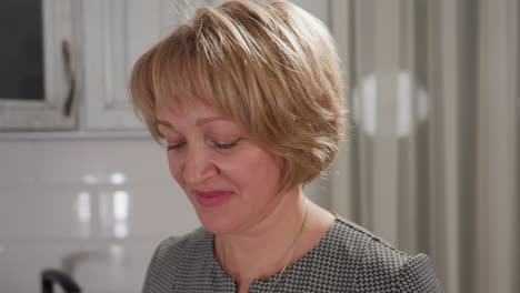 Close up of female artist smiling softly while making gentle body movements as she works joyfully in kitchen with light reflecting on tiled wall and faint curtain patterns in background