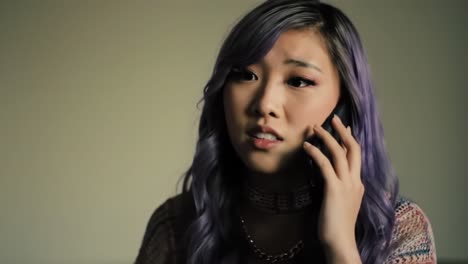 A young woman with purple hair talks on the phone, displaying visible concern and emotion as she listens intently. The background is softly lit, creating a contemplative atmosphere.