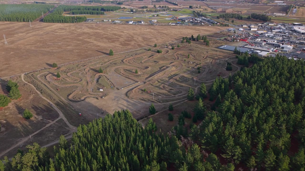 Drone flight over Cromwell's outdoor recreation trails, with dirt tracks, pine trees, and nearby commercial buildings set against the Central Otago landscape.