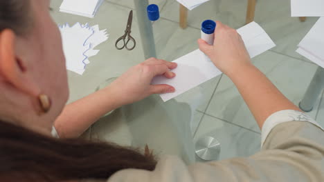 Woman applying glue to paper on glass table in workspace with scissors, folded paper, and craft supplies