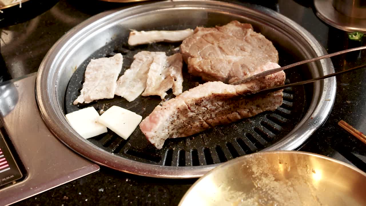 Hand uses tongs to flip pork slices on tabletop Korean barbecue grill under warm lighting