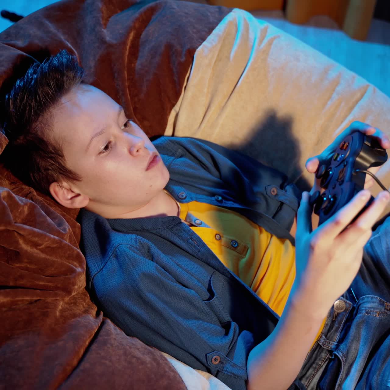 Young gamer playing video games at home. Boy laying in comfortable armchair and managing joystick while playing modern games.