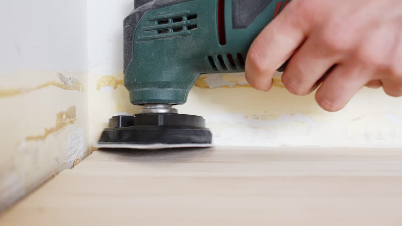 Hand-operated electric sander carefully smooths detailed floor edge areas during interior restoration project showcasing precision equipment technique for quality surface finishing work