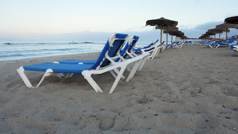 vista lateral de una playa vacía al amanecer con muchas tumbonas y sombrillas de paja, pequeñas olas en el mar, inspiración de viajes de lujo de ensueño